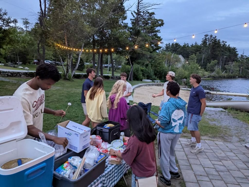 Group of people enjoying a bonfire party outdoors at dusk with string lights overhead.