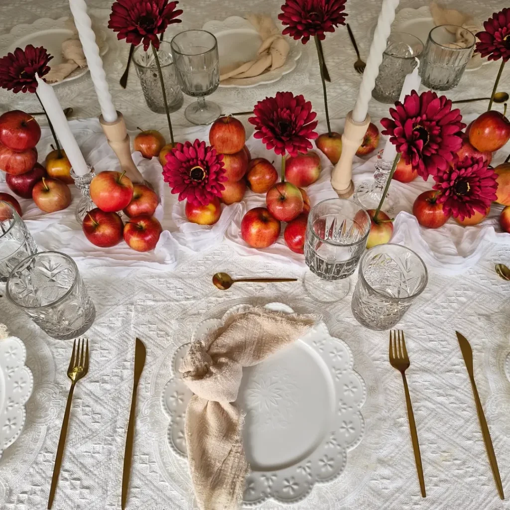 Elegant Thanksgiving table setting with red flowers, apples, candles, and gold cutlery.