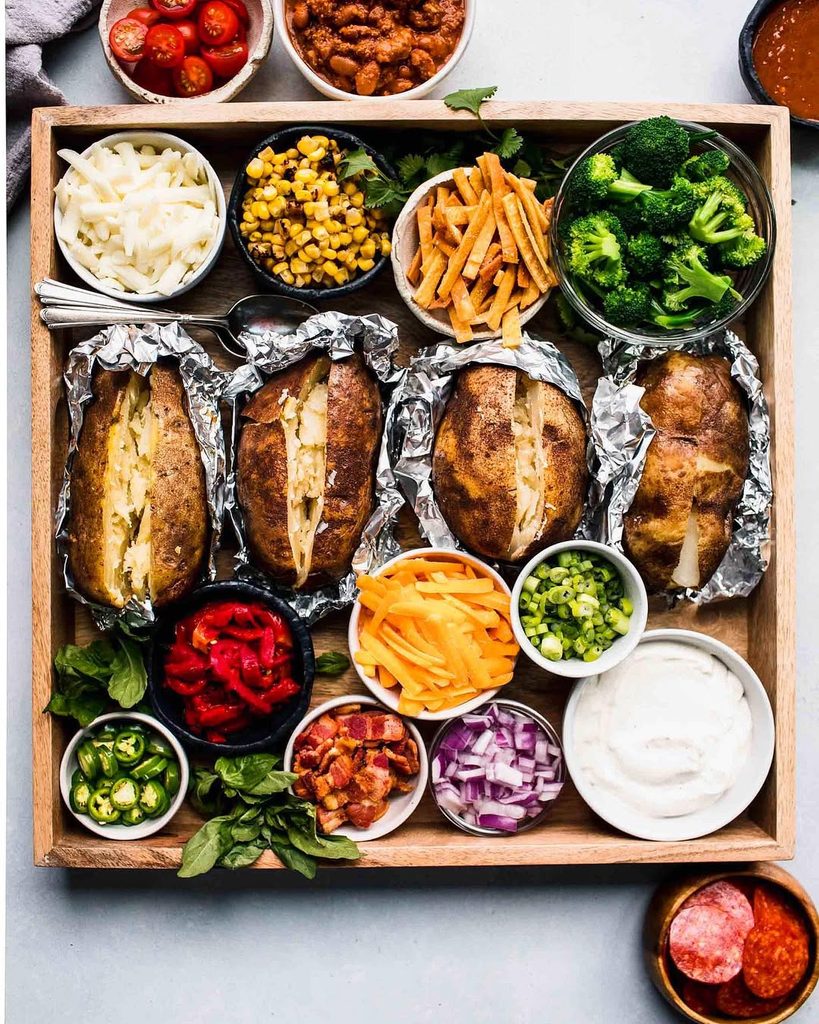 Friendsgiving feast with baked potatoes, broccoli, cheese, and toppings in a wooden tray.