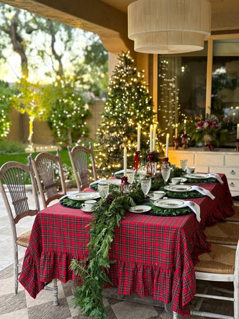 Festive Christmas dining table with red plaid tablecloth, greenery, candles, and decorated tree.