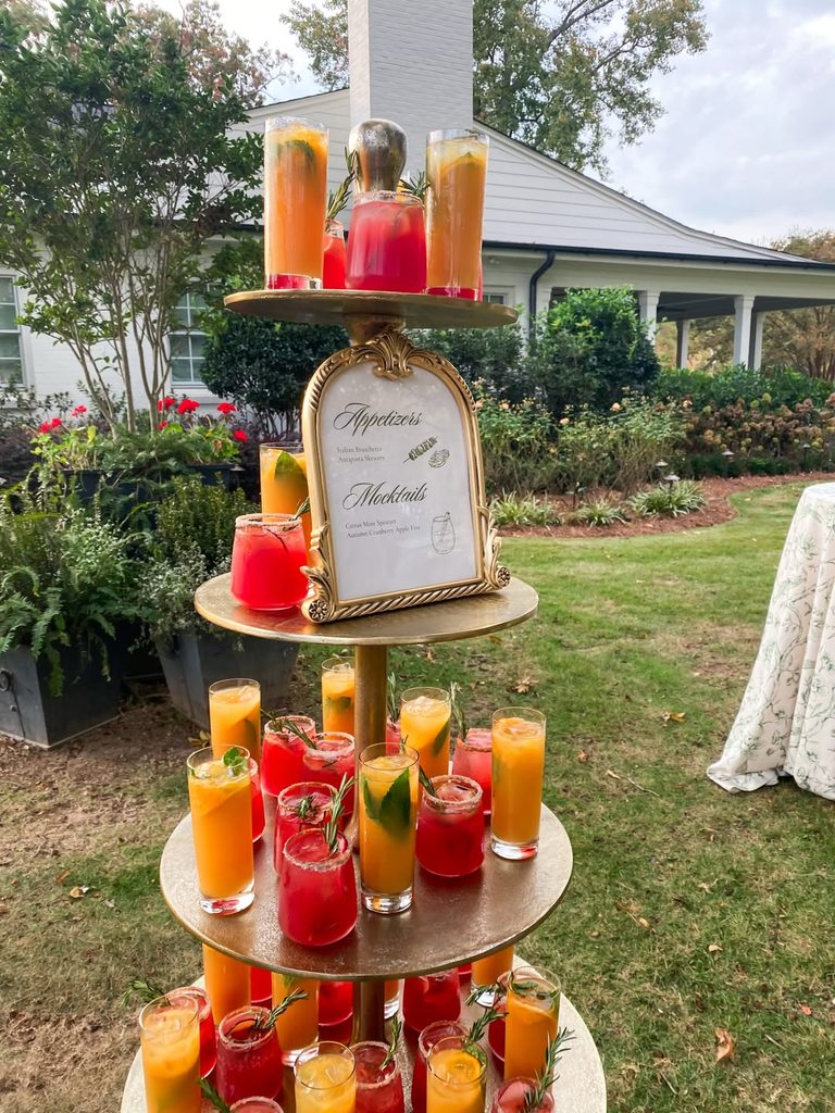 Colorful fall wedding drinks on a tiered stand outdoors with greenery and a house in the background.