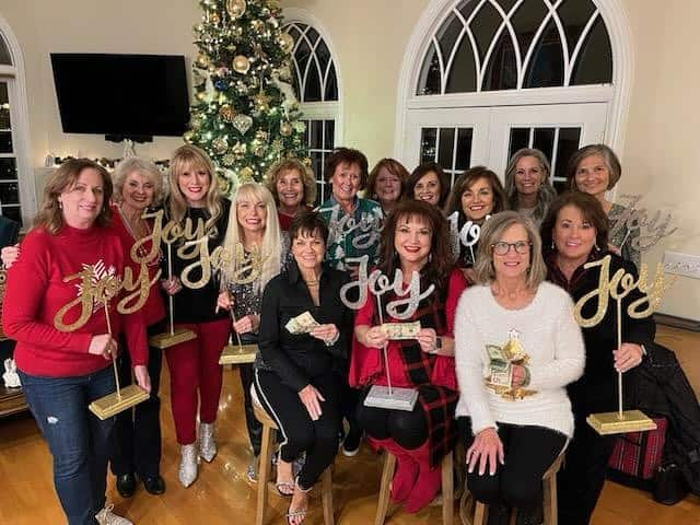 Group of women celebrating Bunco party with Christmas decorations and 'Joy' signs.