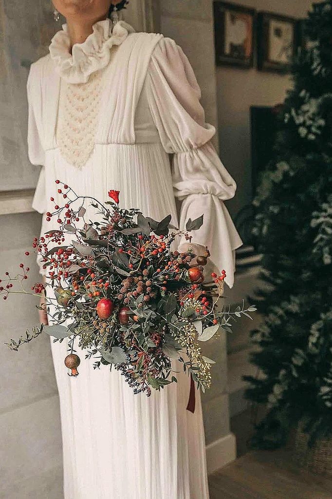 Elegant winter wedding bouquet with red berries and greenery in front of a vintage dress.